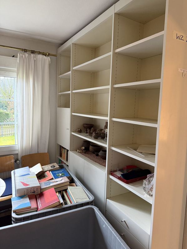Fast ausgeräumtes Bücherregal Ein weißes Regal, in dem nur noch einzelne Gegenstände stehen, davor ein grauer Rollbehälter voller Bücher