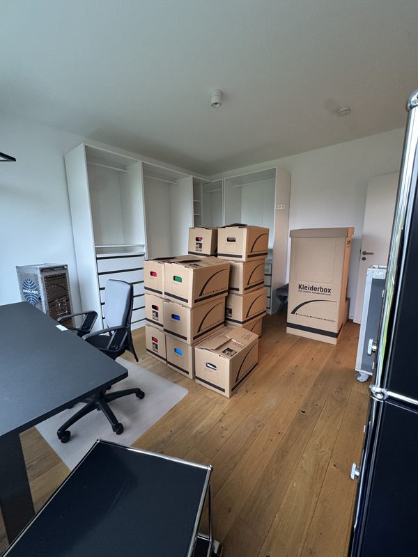 Ein Raum mit großem schwarzem Schreibtisch, Bürostuhl auf einer Bodenschutzmatte und gestapelten Kartons vor einem Einbauschrank