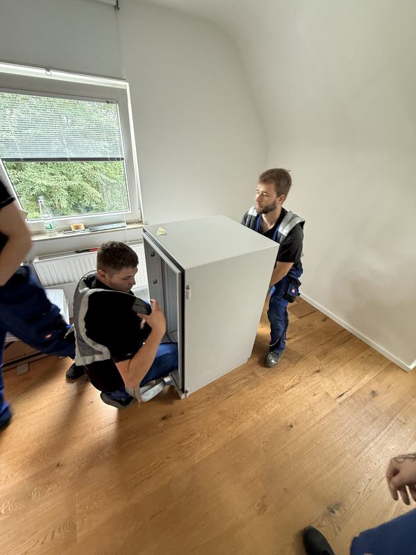 Durch einen leeren Raum tragen zwei Kollegen ein weißes Einbaugerät