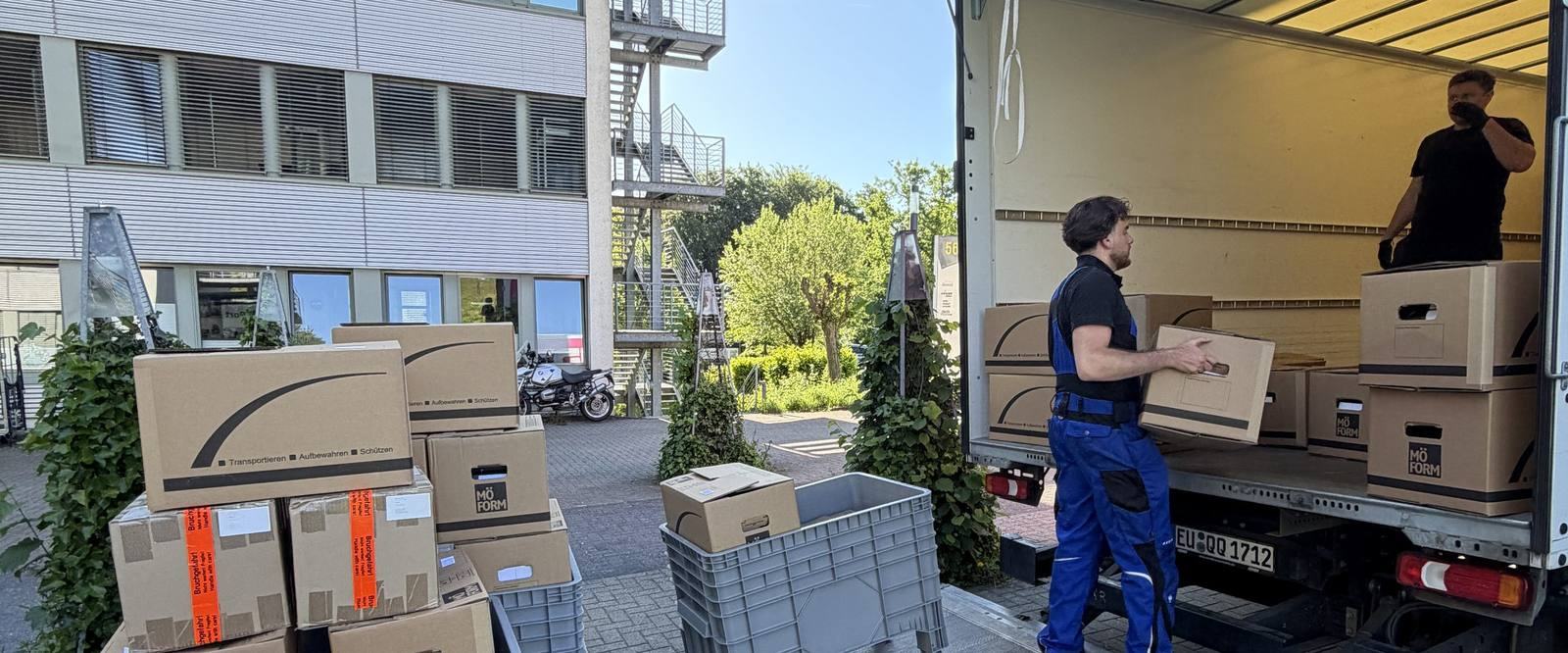 Einblicke in unseren Umzugsalltag in Düsseldorf: Vor einem Bürogebäude mit Außentreppe steht ein Umzugswagen mit offenem Laderaum, ein Mitarbeiter trägt einen Karton zur Ladebordwand, ein zweiter steht im Laderaum, im Vordergrund gestapelte Umzugskartons und graue Kunststoffboxen.