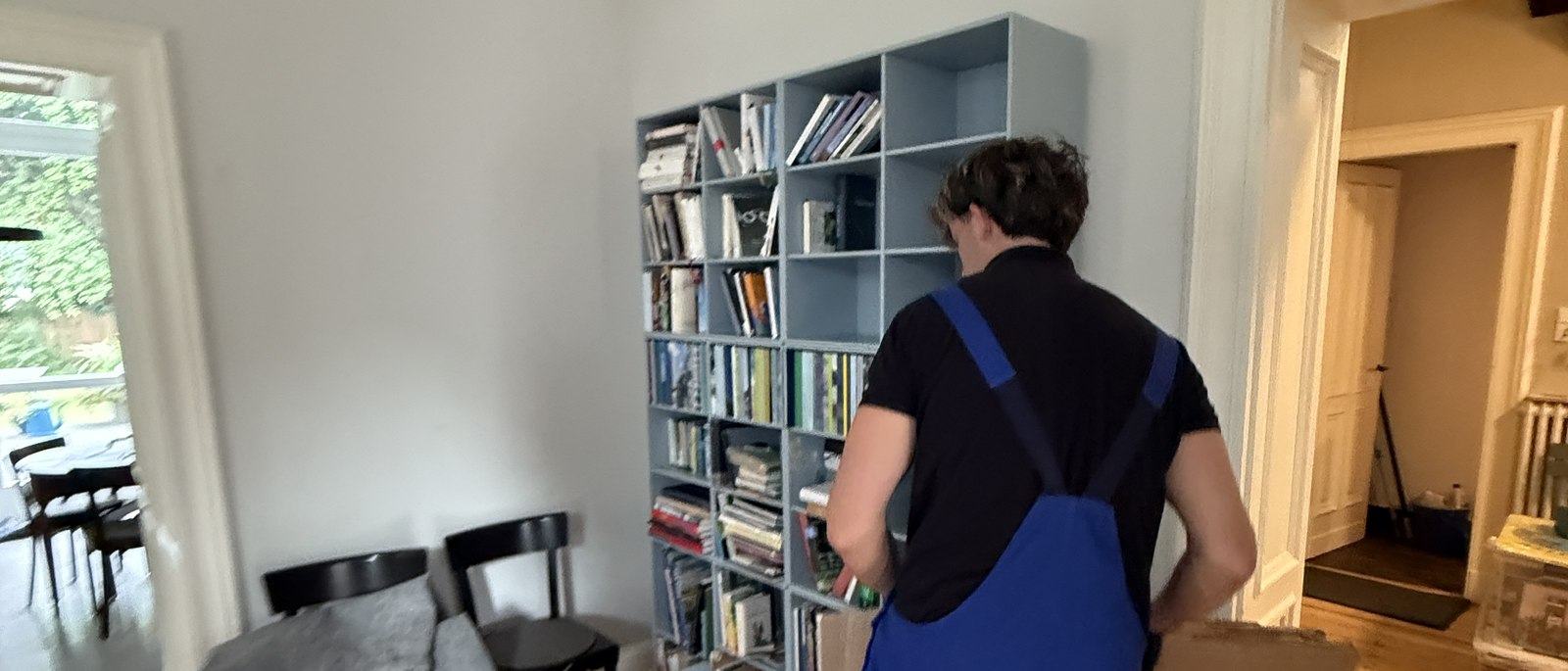Fragen und Antworten zum Umzug in Düsseldorf: Ein Mitarbeiter in Latzhose steht vor einem hellgrauen Würfelregal mit Büchern und Ordnern und packt dessen Inhalt in einen Karton, im Vordergrund ein Esstisch mit Stühlen.