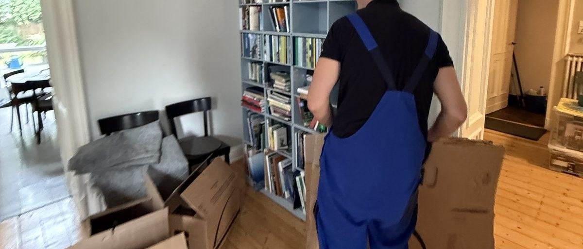 Haushaltsauflösung in Düsseldorf: Ein Mitarbeiter mit Latzhose trägt einen flachen Karton an einem voll bestückten Bücherregal vorbei, davor Stühle und Kartons