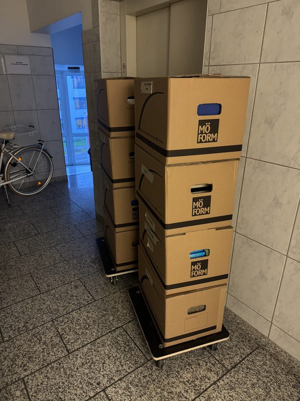 Ein hoher Kartonstapel steht auf einem Rollbrett in einem Flur mit hellem Fliesenboden