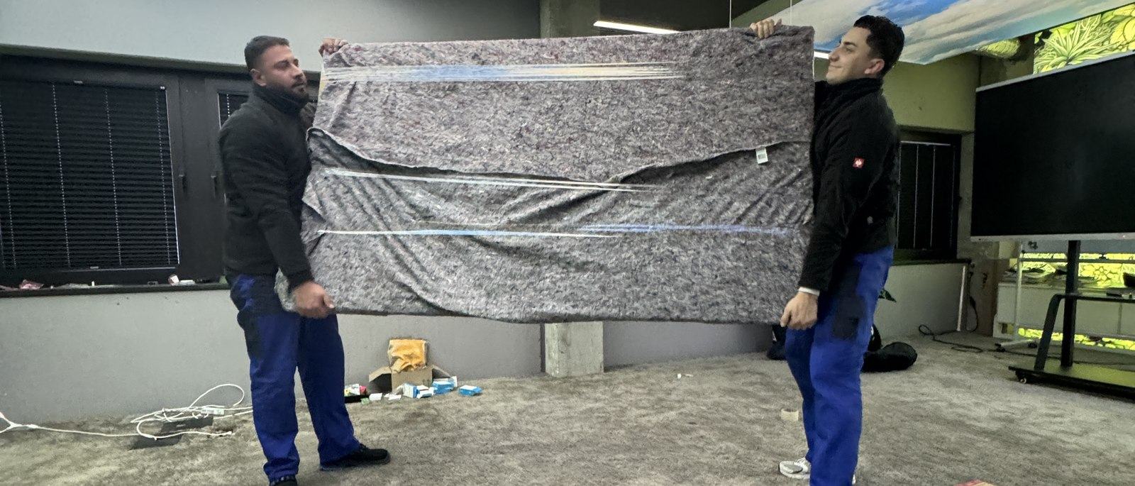 Ihr Kontakt in Düsseldorf: Zwei Mitarbeiter tragen zu zweit ein großes flaches Möbelstück, das in eine graue Umzugsdecke gewickelt ist, über einen Hof an einer Hauswand entlang.
