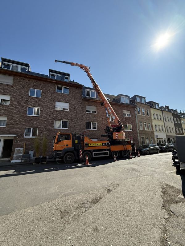 In einer Wohnstraße steht ein orangefarbener Kranwagen, der Ausleger ausgefahren, davor Warnkegel