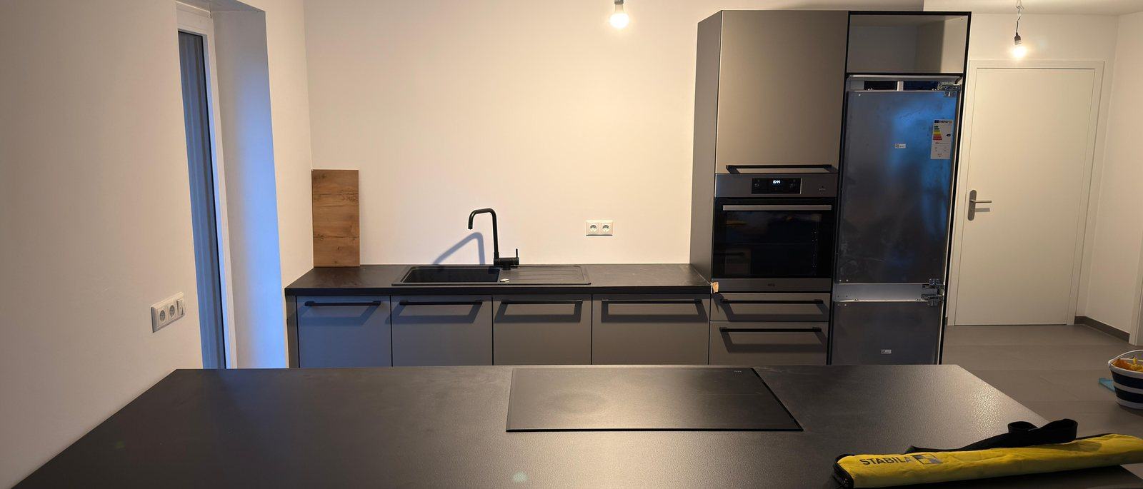 Küchenmontage in Düsseldorf: Eine fertig montierte moderne Küche mit dunklen Fronten, Kochinsel mit Induktionsfeld, Spüle, Backofen und Kühlschrank in einem hellen leeren Raum, auf dem Boden liegt noch eine Werkzeugtasche.