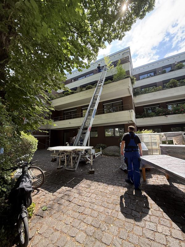 Ein ausgefahrener Möbellift lehnt an einem mehrgeschossigen Wohnhaus, unten wartet ein Mitarbeiter