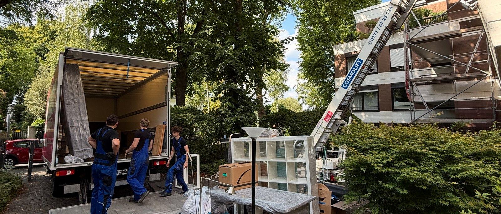 Möbellift für Umzüge in Düsseldorf: Ein ausgefahrener Möbellift ragt bis zum oberen Stockwerk eines eingerüsteten Wohnhauses, davor drei Mitarbeiter an der offenen Ladefläche eines Lkw sowie ein Regal und Kartons