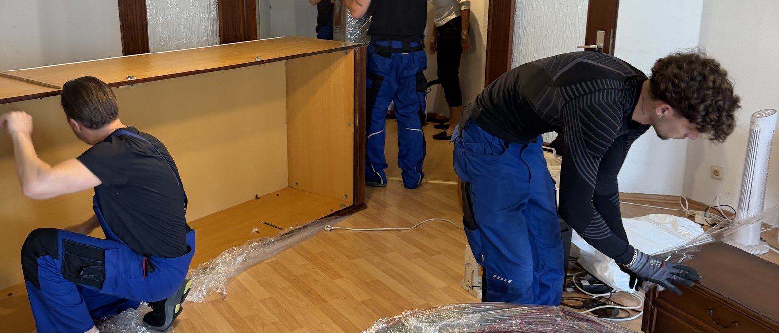 Möbelmontage in Düsseldorf: Drei Mitarbeiter zerlegen in einem Wohnzimmer mit Parkett eine große Schrankwand aus Holz, die Seitenteile liegen bereits am Boden, daneben Folie und Werkzeug.