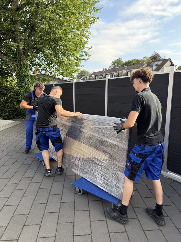 Auf Rollbrettern schieben drei Kollegen ein foliertes Möbelstück über eine Einfahrt