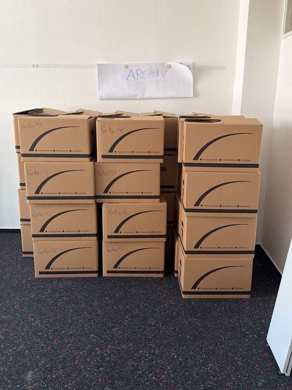 In einem leeren Büroraum stapeln sich nummerierte Umzugskartons unter einem von Hand geschriebenen Schild