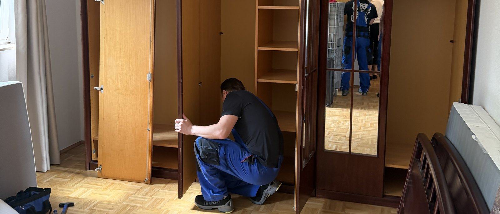 Seniorenumzug in Düsseldorf: Ein Mitarbeiter hockt vor einem leergeräumten dunklen Holzschrank, das Schlafzimmer hat Parkettboden