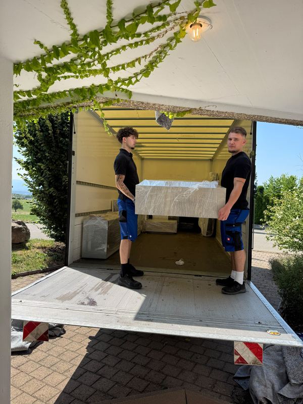 Zwei Mitarbeiter hieven ein folienverpacktes Möbelstück über die abgesenkte Ladebordwand hinein in den Laderaum des Umzugswagens