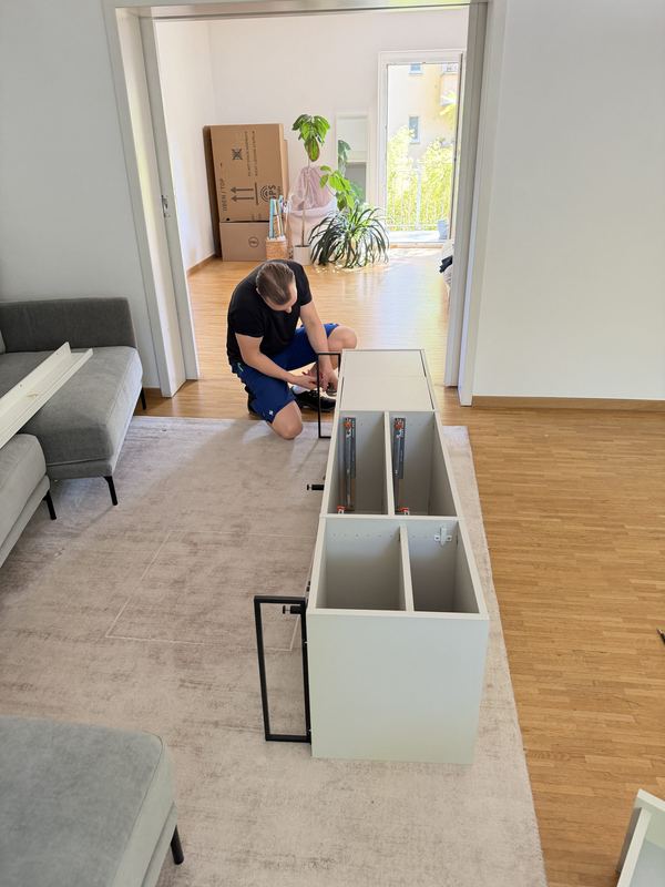 Ein kniender Mitarbeiter baut in einem hellen Wohnzimmer ein weißes Sideboard zusammen, dahinter stehen Umzugskartons