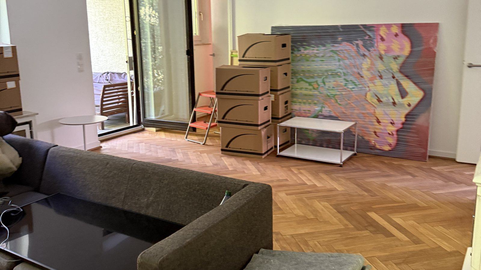 Umzugscheckliste für Düsseldorf: In klaren Schritten ins neue Zuhause: In einem Wohnzimmer mit Fischgrätparkett stehen gestapelte Umzugskartons neben einem Sofa und einem Beistelltisch, an der Wand lehnt ein großes buntes Bild, dahinter eine Terrassentür.