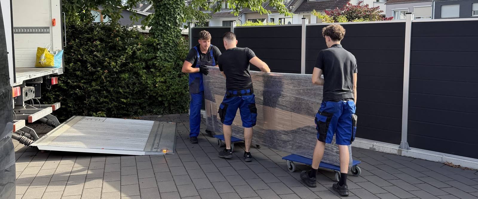 Umzugsunternehmen Düsseldorf: Drei Mitarbeiter bringen ein foliertes Möbelstück auf Rollbrettern über einen Hof zum Umzugswagen mit abgesenkter Ladebordwand