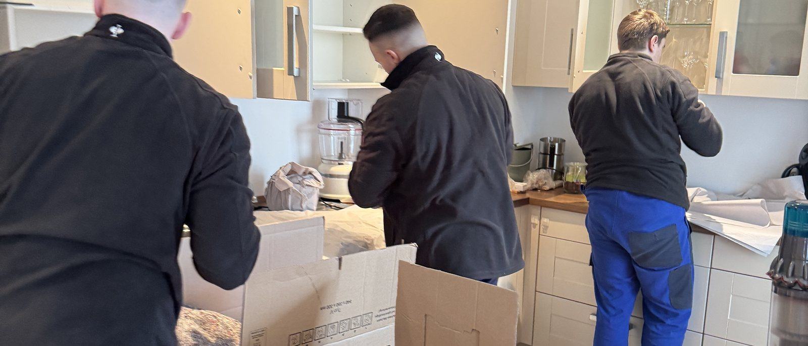 Umzugskartons und Kleiderboxen für Ihren Umzug in Düsseldorf: Drei Mitarbeiter packen in einer Küche mit hellen Fronten Geschirr und Kleingeräte in Umzugskartons auf der Arbeitsplatte, in der Mitte ein offener Karton mit Packpapier.