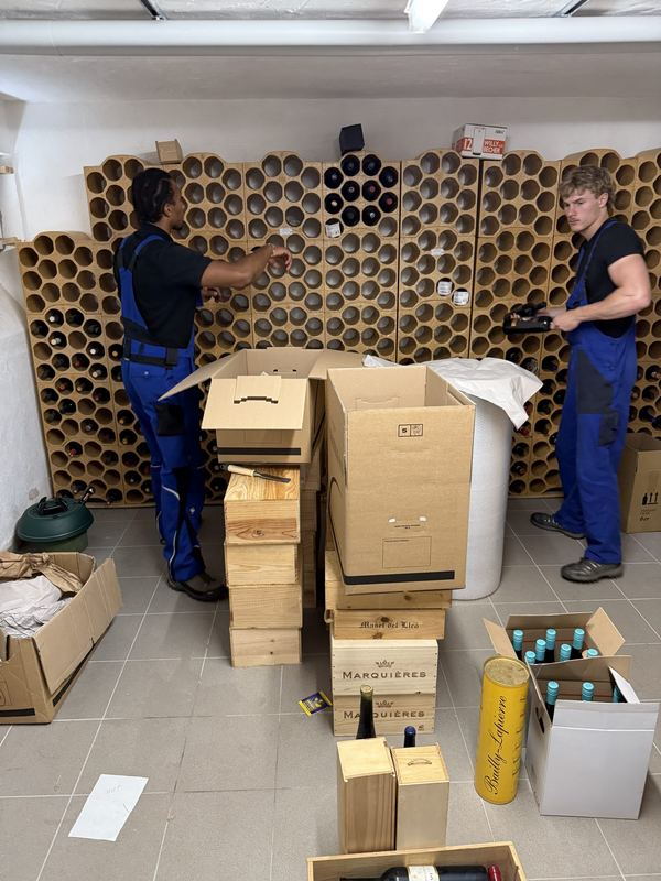 Zwei Mitarbeiter räumen Weinflaschen aus dem Tonröhrenregal eines Kellers und verstauen sie in Holzkisten und Kartons