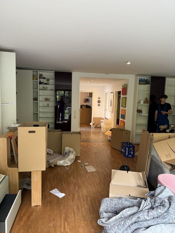 Ein Wohnzimmer mit schon leeren Einbauregalen und bereitgestellten Kartons, im Hintergrund zwei Kollegen